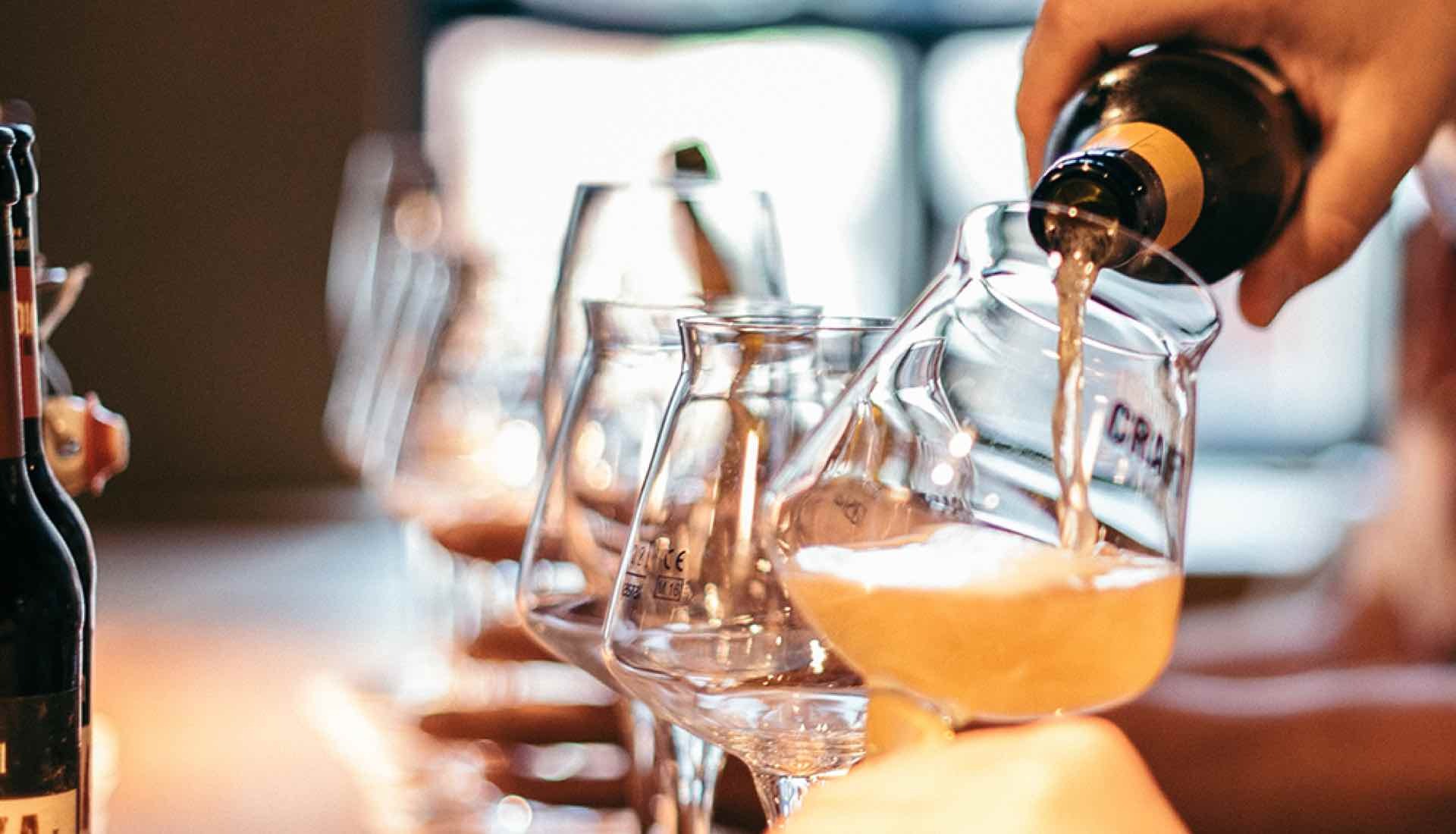 Nahaufnahme von einer Hand, die Bier aus einer Flasche in ein Glas einschenkt; mehrere leere Gläser stehen daneben auf einem Tisch.