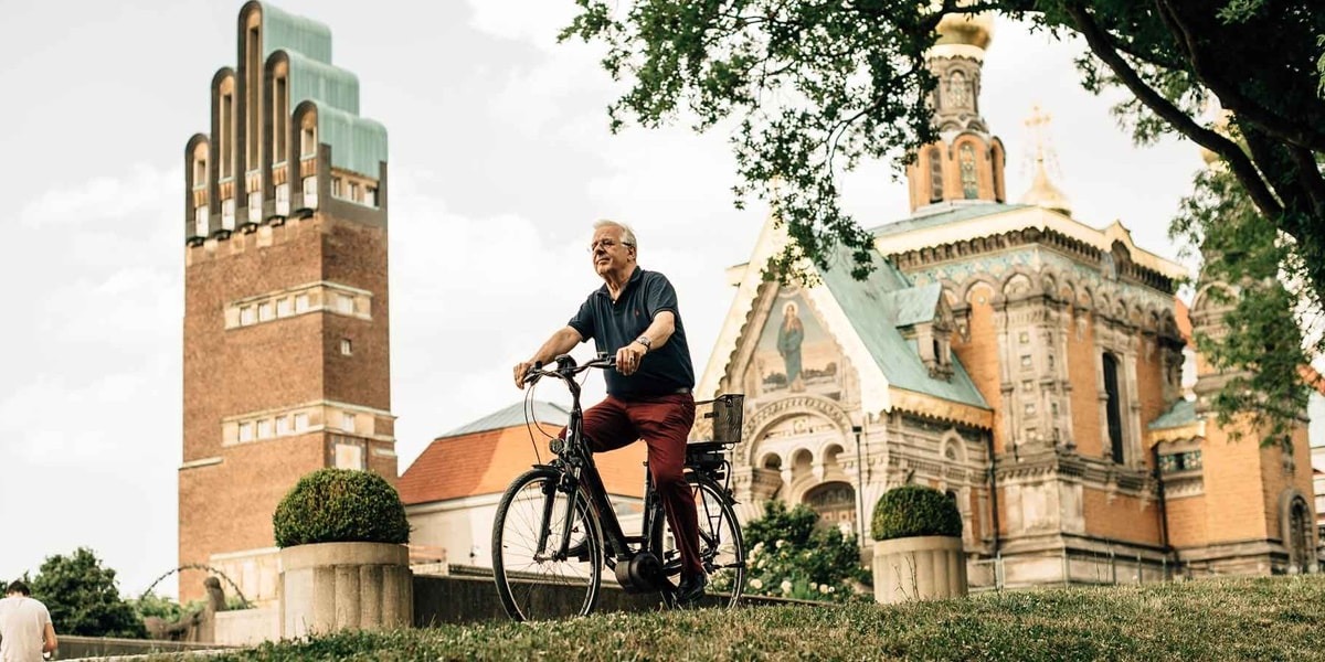 Älterer Mann fährt Fahrrad vor der russischen Kapelle und dem Hochzeitsturm auf der Mathildenhöhe in Darmstadt, umgeben von Bäumen und Rasen.