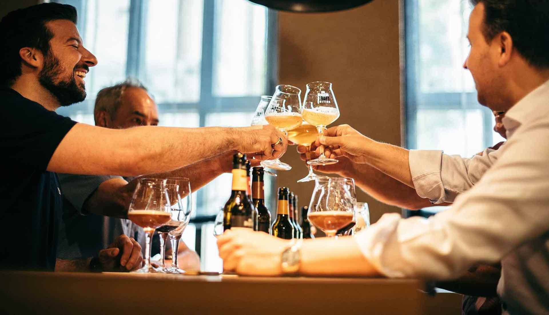 Vier Männer sitzen an einem Tisch, stoßen mit Craft-Biergläsern an und lachen. Auf dem Tisch stehen mehrere Bierflaschen und Gläser. Im Hintergrund sind große Fenster.