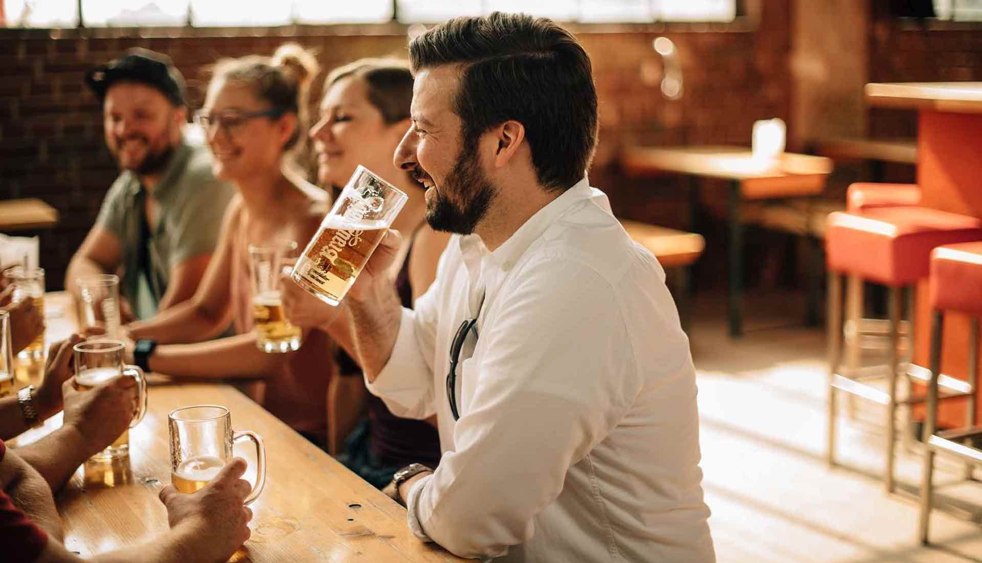 Mehrere Personen sitzen lachend an einem Holztisch in einer Bar und trinken Bier aus Gläsern.