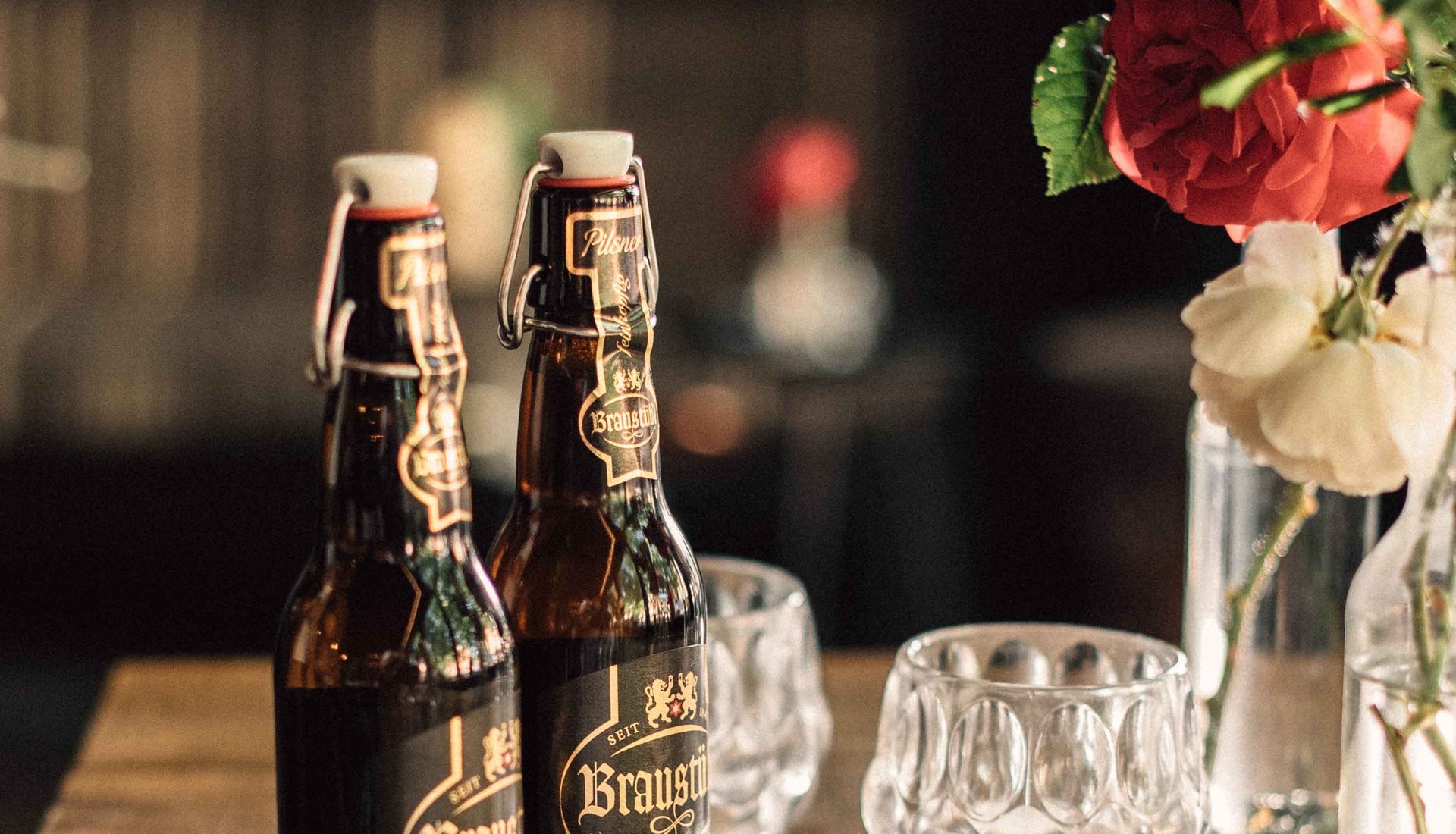 Zwei Flaschen Pilsner Bier mit Bügelverschluss auf einem Holztisch, daneben zwei leere Biergläser und eine Vase mit roten und weißen Blumen.
