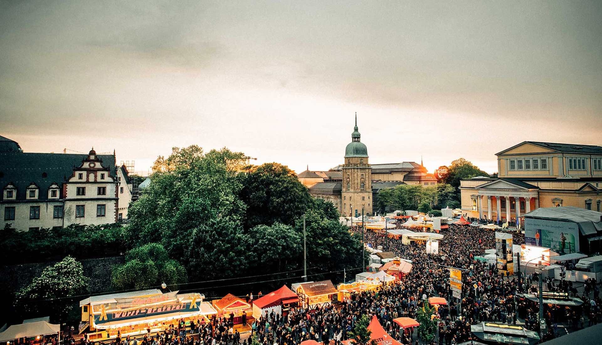 Großes Stadtfest mit Menschenmenge, bunten Marktständen und historischen Gebäuden im Hintergrund bei Sonnenuntergang.