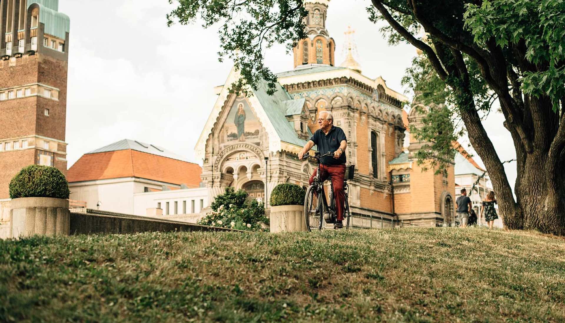 Ein älterer Mann fährt Fahrrad auf einer Wiese vor einer historischen Kirche mit verzierten Fassaden; im Hintergrund Bäume und weitere Personen.